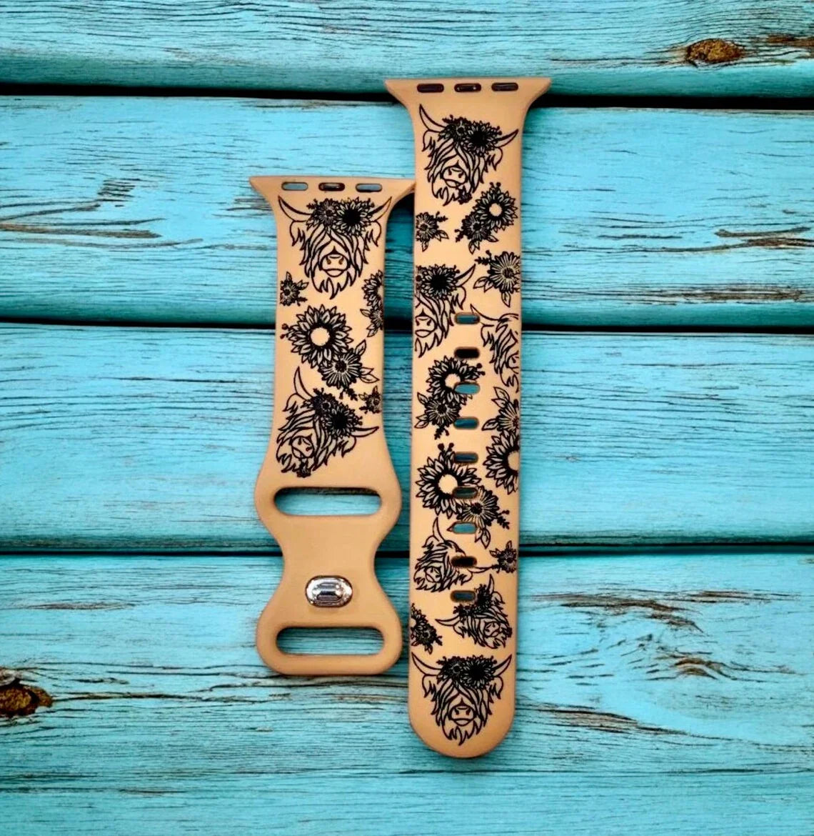 Beige Apple Watch straps with black floral and cow designs on teal wooden background