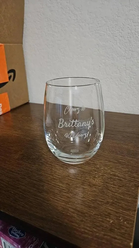 Personalized stemless wine glass engraved with 'Cheers to Brittany's 40 years!' on wood shelf