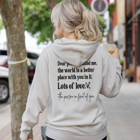 Woman in light hoodie with inspirational text on back holding iced coffee walking on city sidewalk