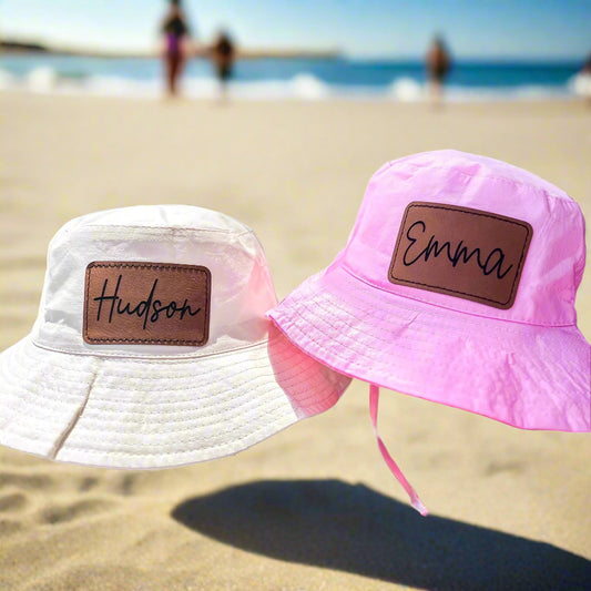 Two bucket hats, one white and one pink, with brown patches displaying names.