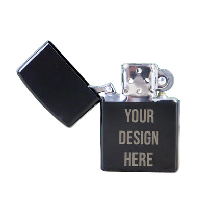 Black Zippo-style lighter with 'Your Design Here' text on a light wood surface with green leaves in the background.