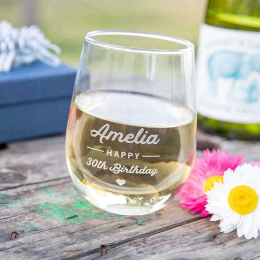 Stemless wine glass with engraved text on a wooden surface with flowers and a bottle in the background.